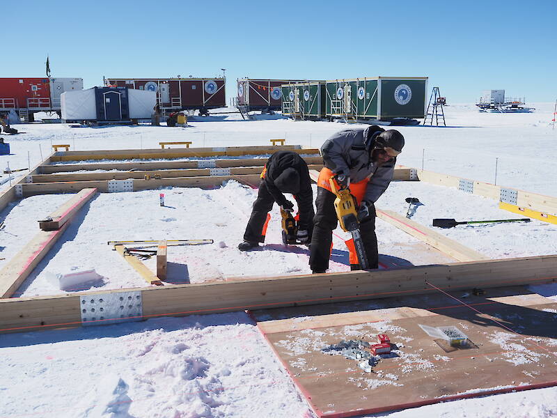 Two men construct a wooden frame in the snow, with shipping container vans that provide accommodation and mess facilities for the campsite in the distance.