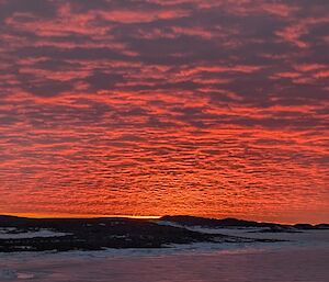 The sun sets behind a distant hill from Davis Station producing a vibrant sunset, the sky is glowing pink and orange.