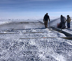 Five people use shovels to compact snow being blown in to a wooden frame