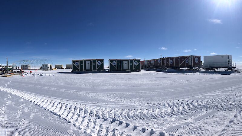 A panoramic view of shipping containers and a drill shelter under construction, with blue sky and a vast expanse of white ice.