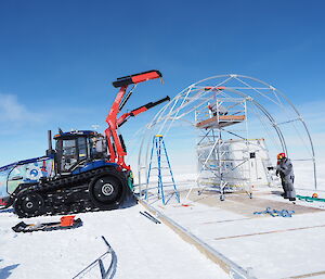 Cranes on the back of a tractor help position a structure inside a metal tent frame.
