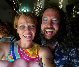 A woman in a multi-coloured top and a man with a beard wearing eye make up