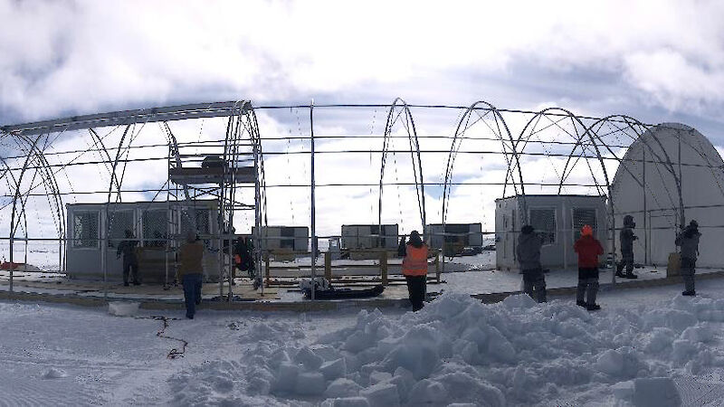 People standing on one side of a large, cylindrical tent frame, preparing to pull a cover over the top using ropes.