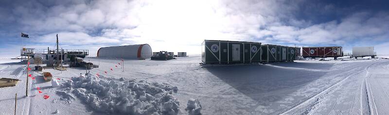 A collection of red and green shipping containers and a large cylindrical tent, on a flat, white, ice plateau