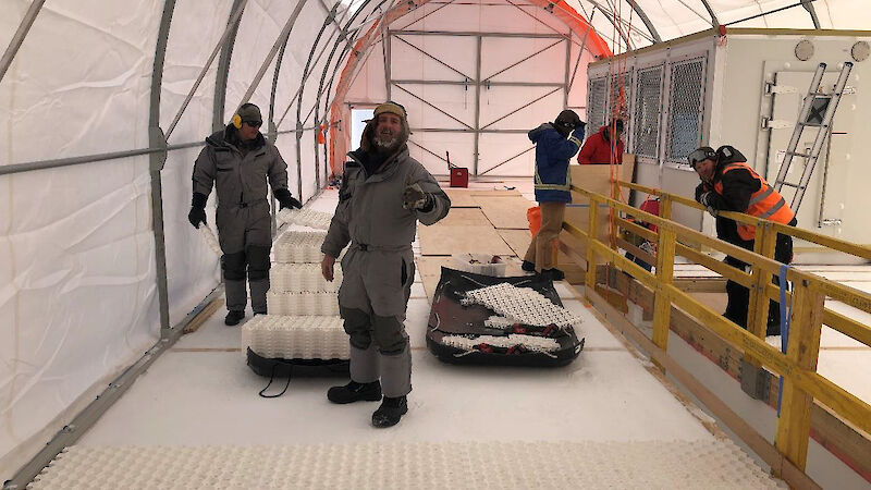 Two people laying gridded, white, plastic tiles over compacted snow, inside a large tent.
