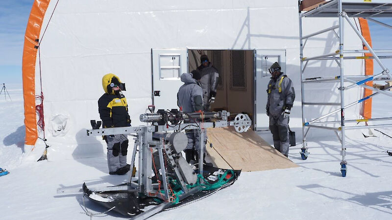 An ice core drill on a sled being moved up a wooden ramp into a large, semi-circular tent.