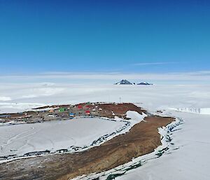 Distant image of Mawson Station. In the foreground image displays ice and rock. The mid-ground features the Mawson station colours buildings dotted across a band of brown rock. In the distant you can see snow to the horizon with a mountain in the center of the image.