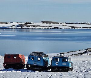 Image focuses on a Hagglund vehicle towing a caboose. The background shows a series of rocky hills covered in snow. In the centre of the image there is a deep blue body of water.