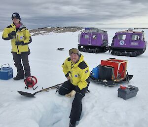 Focus of the image is an expeditioner dressed in yellow survival clothing kneeling beside a hole in the snow with equipment scattered around. The sky is grey and cloudy and reaches to the horizon line. Along the horizon line is a rocky hill.