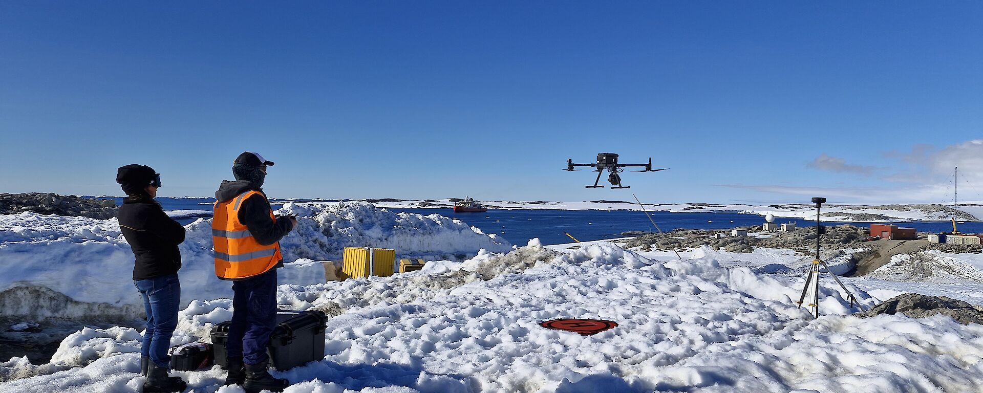 Image depicts two people standing to the left of the image with a drone hovering at eye-level to the right of the image. The ground is snow with equipment scattered throughout the image. The background features a body of water with snow covered hills in the distance.