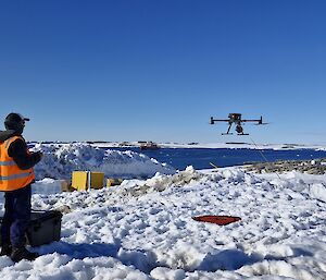 Image depicts two people standing to the left of the image with a drone hovering at eye-level to the right of the image. The ground is snow with equipment scattered throughout the image. The background features a body of water with snow covered hills in the distance.