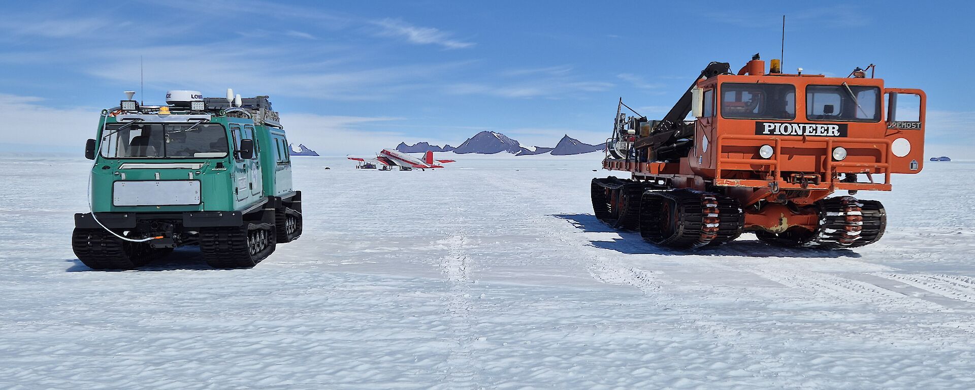 A green snow tractor on the left and an orange one on the right, on a flat snowy plain