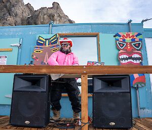 A man stands on the deck of a shed, wearing a pink top and a red and white cowboy hat, with two big speakers.