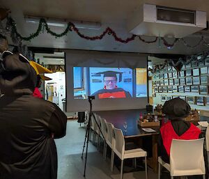 Person in graduation robes sits as large table, watching projector screen. On screen is a professor in robes.