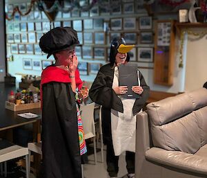Graduate stands in robes and oversized bonnet, while presenter is in penguin costume and holds clip board