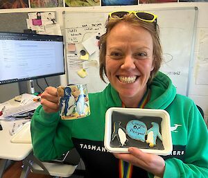 Person sitting at computer desk, smiles to camera and holds up a mug, with little penguin motif, and penguin cookies
