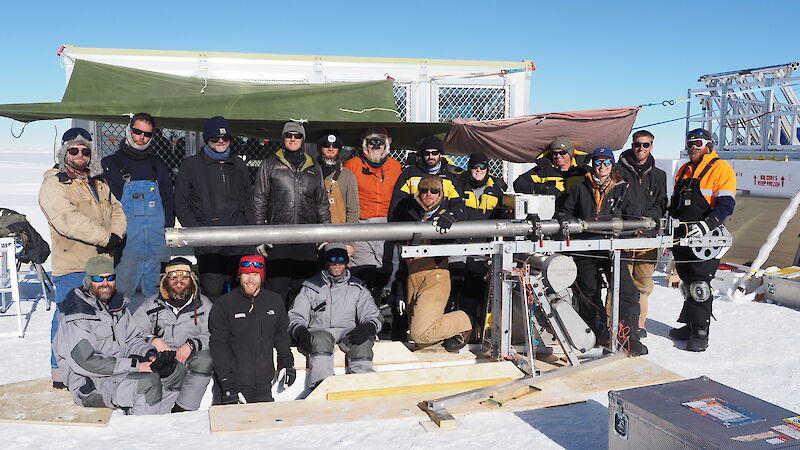 A group of people posed around a horizontal drill barrel lifted about one metre off the snow surface.