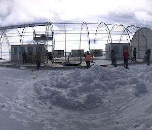 Metal framework for a semi-circular tent, with people standing along one side holding ropes to pull a cover over the top