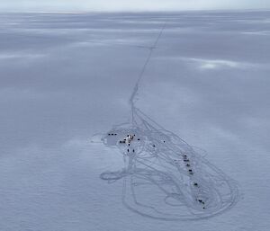 Aerial view of the ice plateau showing a line of tractors and a small camp area surrounded by vehicle tracks.