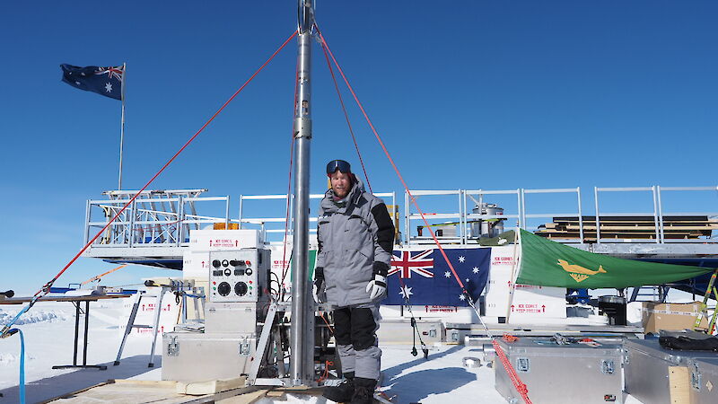 A man standing beside a vertical metal pipe that is tied down with three ropes and suspended over a trench in the snow