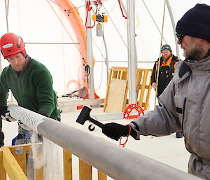 One man removes an ice core from a horizontal metal drill barrel while a second man taps the drill barrel with a small plastic hammer