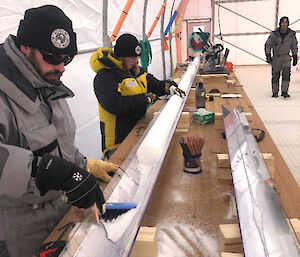 Two people use a small brush to remove loose ice from an ice core sitting in a v-shaped channel on a table