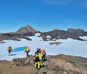 Six expeditioners walking over big rocks with a snowy slope in the background
