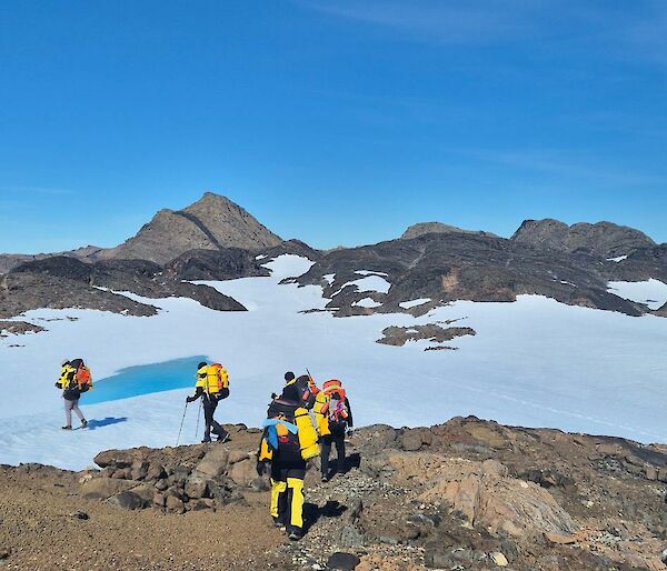 Six expeditioners walking over big rocks with a snowy slope in the background