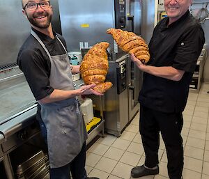 Two chefs hold up huge croissants in a kitchen