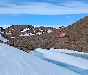 A red melon hut on the side of a rocky hill with snow in the foreground