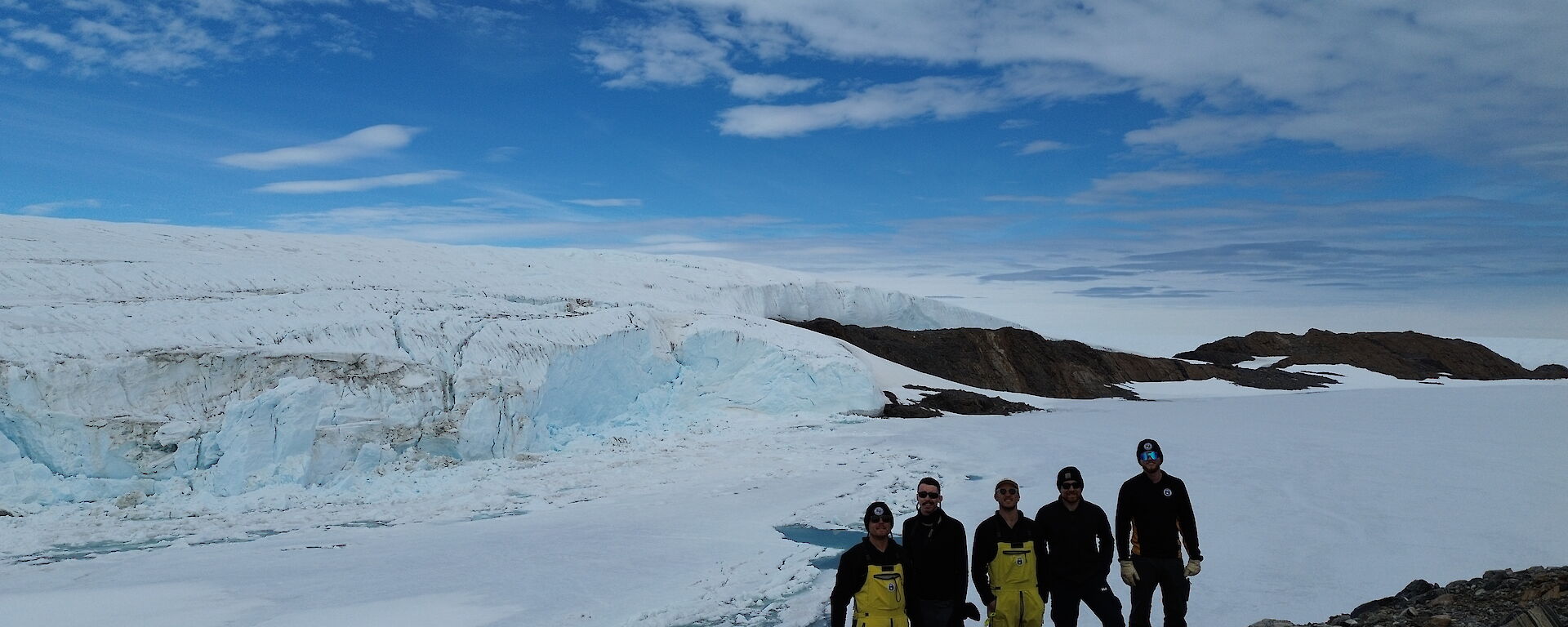Wide shot of five people against a snowy background, with a blue glinting glacier