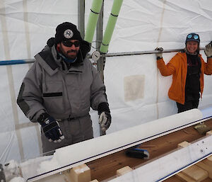 A man and woman standing beside an ice core sitting in a v-shaped channel on a wooden table