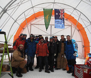 A group of 15 people standing inside a semi-circular tent, wearing warm clothing.