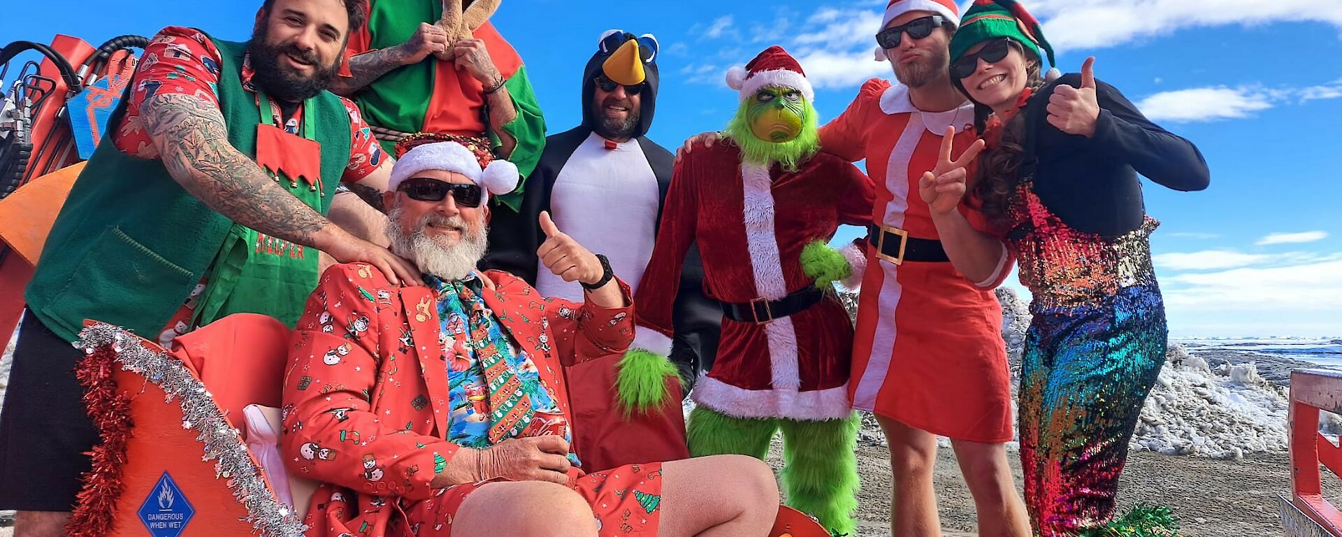Seven AAD expeditioners are dressed in Christmas costume. One Dressed as 'Santa' sits in a red sleigh.