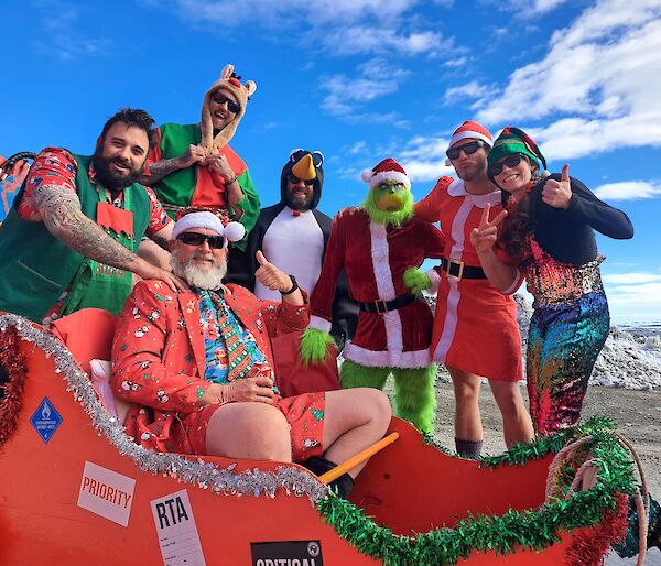 Seven AAD expeditioners are dressed in Christmas costume. One Dressed as 'Santa' sits in a red sleigh.
