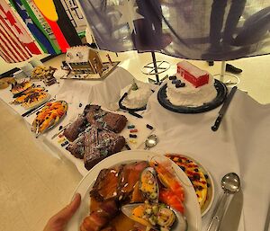 Image is of plate of food served at Christmas lunch - Muscles, Prawns, meat are visible. The background features the table spread of deserts.