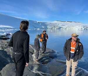 4 Expeditioners in high-vis work ware stand on rocks adjacent to bay of water. A Karge Ice cliff fills the mid-ground and a blue sky fills the backdrop.