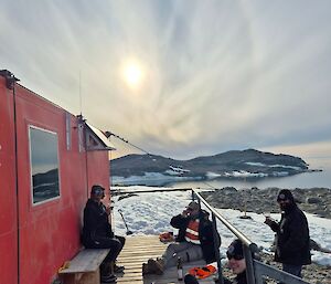 Image of four expeditioners sitting on deck at front of Robbo's Hut. Robbo's Hut is a small red portable building that fills the left half of the image. The sky shows a sun with a halo and veil of cirrus clouds. In the mid area of the image a hill of brown rocks dotted with snow is visible.