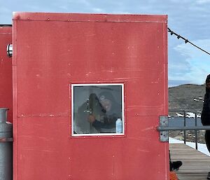 Image shows one expeditioner through a window in the side of Robbo's Hut (Red portable building) sitting down (on the toilet) with another expeditioner outside (to the right of the image) on the deck.