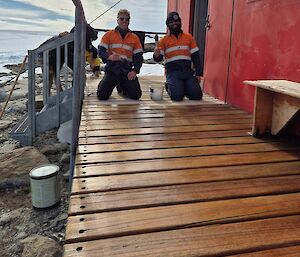 Image shows two carpenters kneeling on the deck at the front of Robbo's Hut (red portable building) holding paintbrushes and smiling (while they are oiling the deck). Foreground shows freshly oiled timber deck. Background shows snowy landscape and cloudy sky.