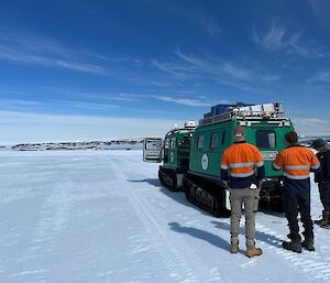 Image features three trades people standing behind a Green Hagglund Vehicle. Foreground displays the firm snow terrain marked by Hagglund tracks. The background shows blue sky and rock features.