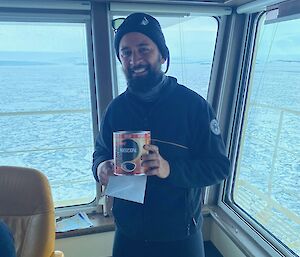 A smiling bearded man in black polar gear and a black beanie holding an instant coffee tin