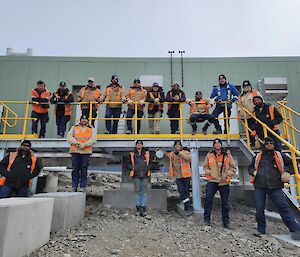 A group of people in hi-vis stand on a yellow deck in front of a green metal building