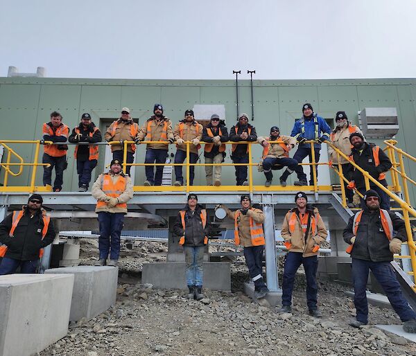 A group of people in hi-vis stand on a yellow deck in front of a green metal building