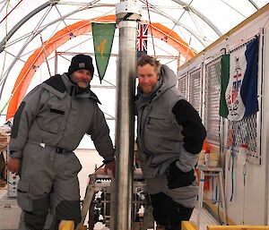 Two men stand beside a large metal cylinder inside a drilling tent