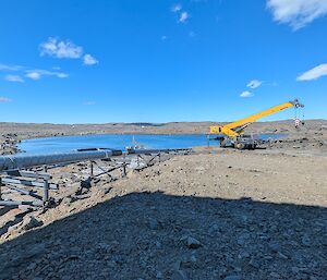 View over gravel towards a body of water with a yellow crane parked near the water's edge