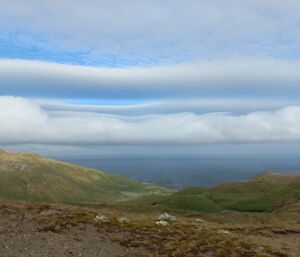 A puffy long, horizontal white cloud that looks like it's sitting on glass, above green rugged hills