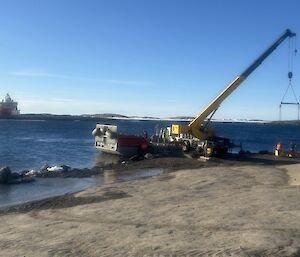 A crane and barge and the water’s edge, with the Nuyina in the background