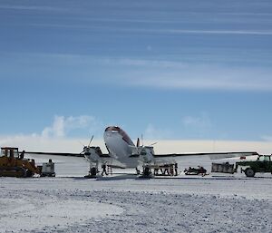 Image focus is of the DC3 aircraft. Adjacent to the aircraft are tractors and equipment. The background features a bright blue sky with a snowy ground.
