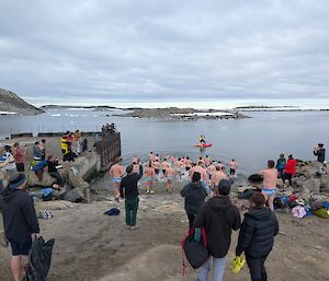 Image of a large group of people in bathers entering the water from the Casey Boat ramp. The mid ground shows the ocean water inside Newcomb Bay with one person on a rescue canoe vessel. The background features rocky hills and islands with a cloudy sky.